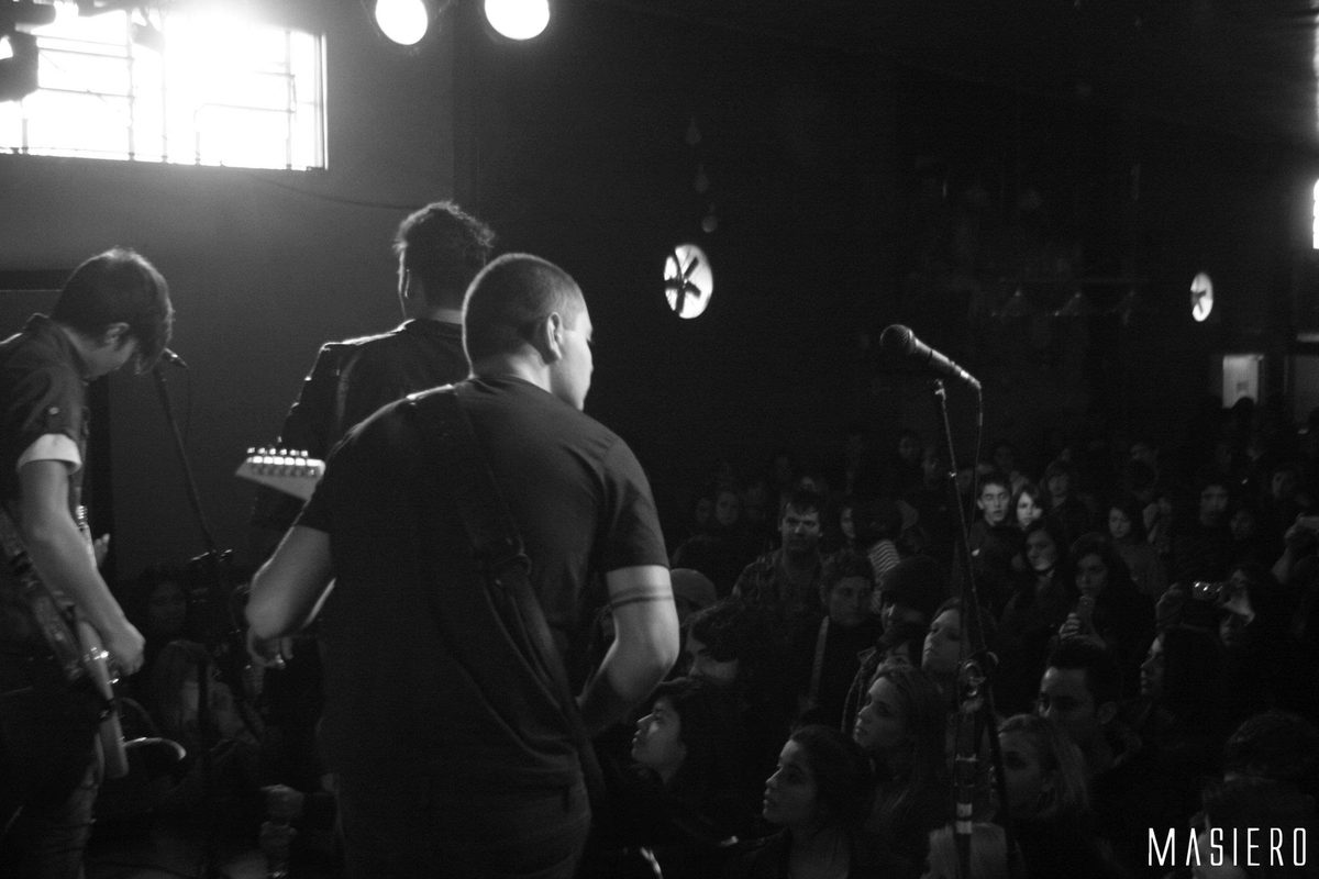 Banda no palco vista de trás, com o público iluminado, em preto e branco.