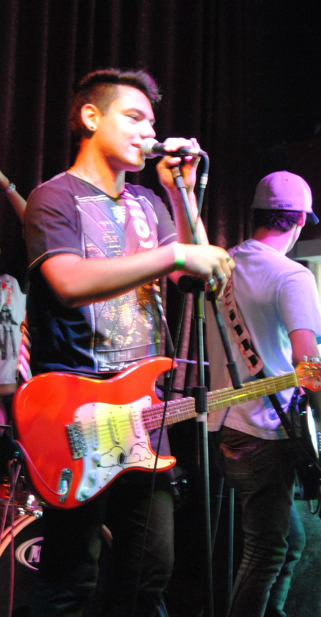 Vocalista com guitarra vermelha cantando no palco, cortina vinho ao fundo.